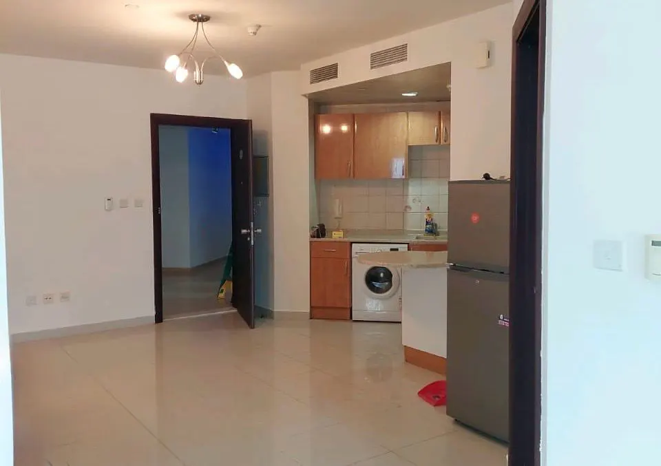 A bright, empty apartment with tile floors, an open kitchen with wood cabinets, and a stainless steel refrigerator.