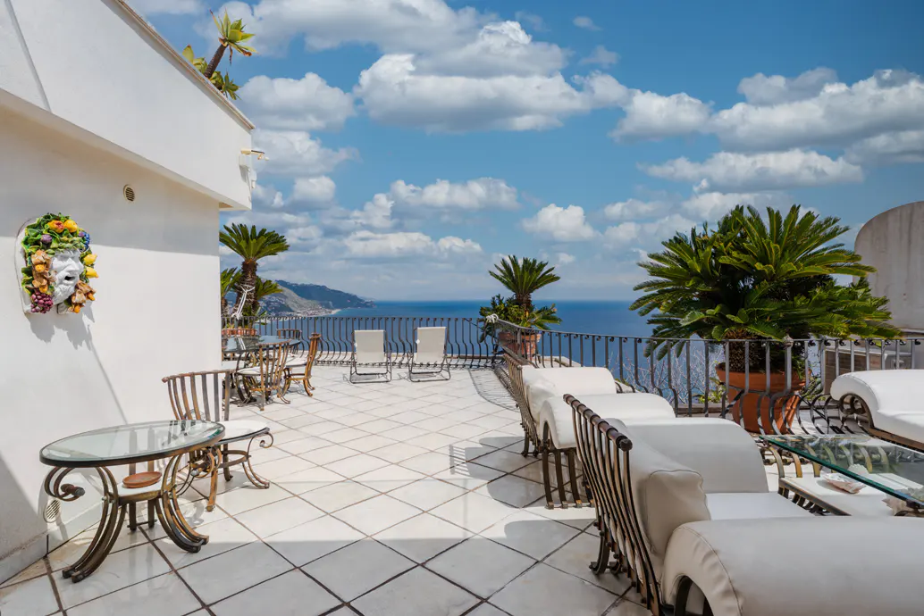 Outdoor patio with white tile, lounge chairs, tables, and ocean view under a blue sky with white clouds.