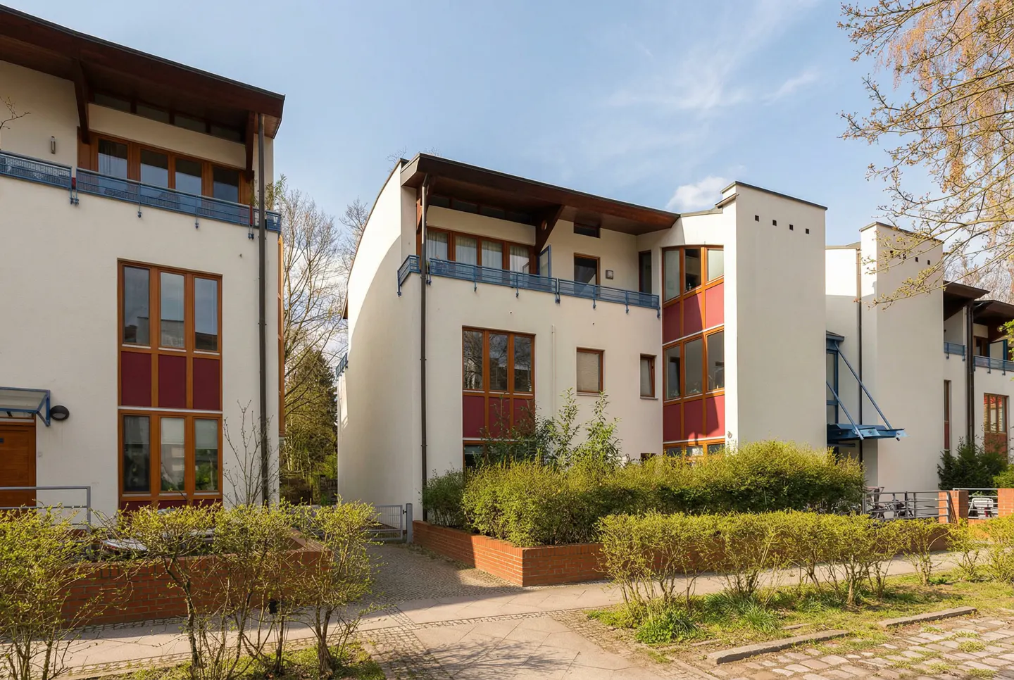 Modern white townhouses with red accents, brown trim, and blue balconies, surrounded by green bushes under a blue sky.