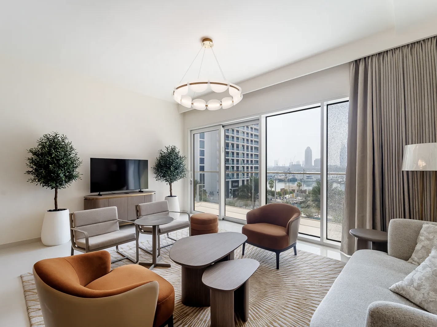Bright living room with a TV, sofa, chairs, and a view of the city skyline through a large window.