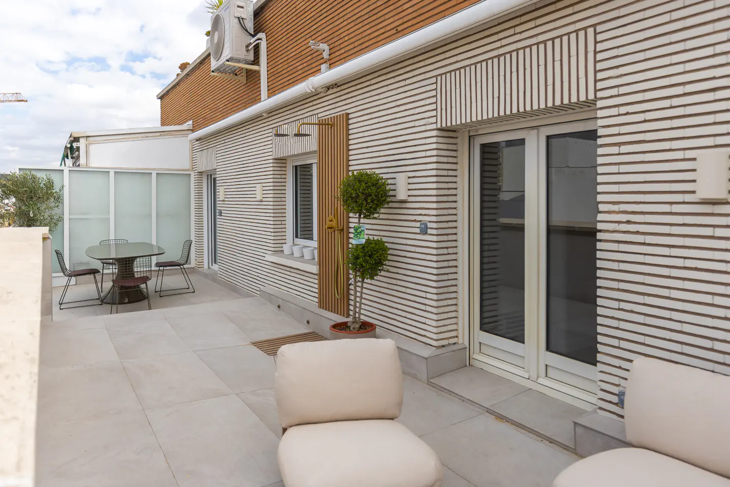 A rooftop patio with a table, chairs, and lounge seating. The building has a brick facade with white trim and a small tree in a pot.