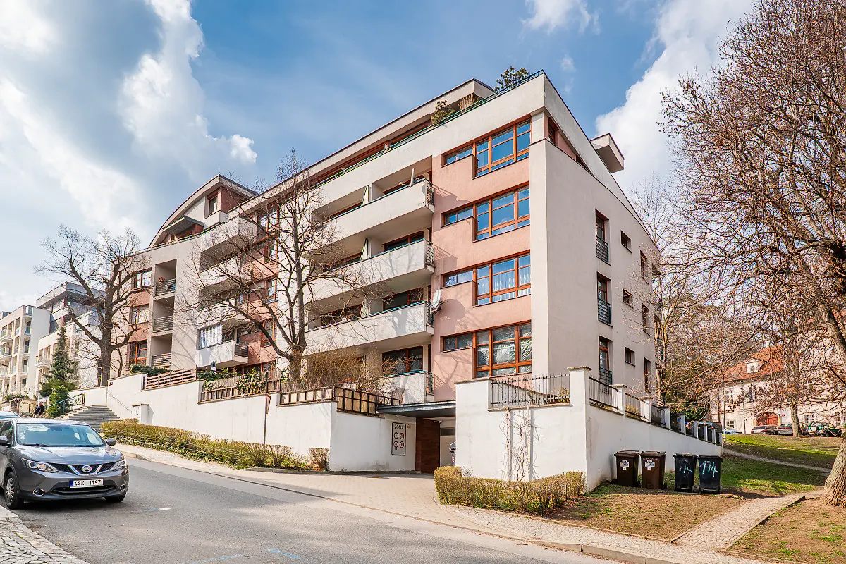 Modern apartment building with balconies, painted in light pink and white, on a sunny day with blue sky and trees. A car is parked on the street.