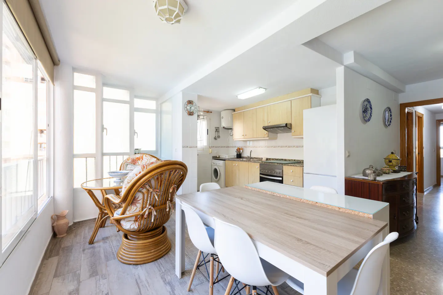 Bright, open-concept kitchen and dining area with light wood table, white chairs, and rattan furniture near large windows.