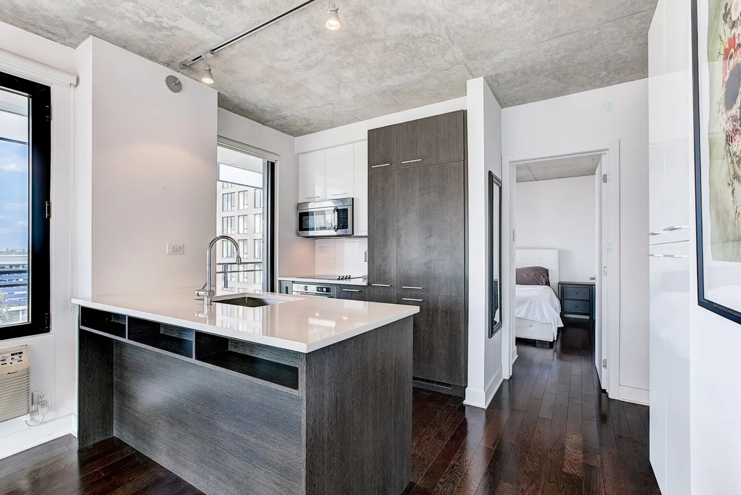 A modern kitchen with a gray island, white countertops, and stainless steel appliances. A bedroom is visible through a doorway. Dark wood floors.