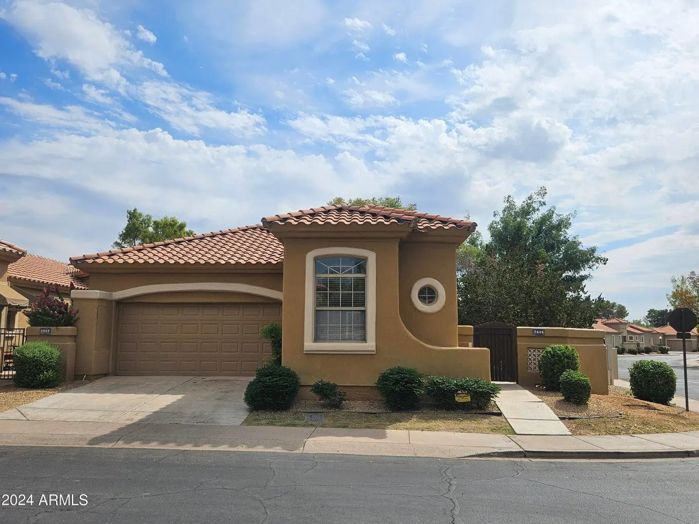 Tan single-story home with a red tile roof, a two-car garage, and green bushes in front. Blue sky with white clouds above.