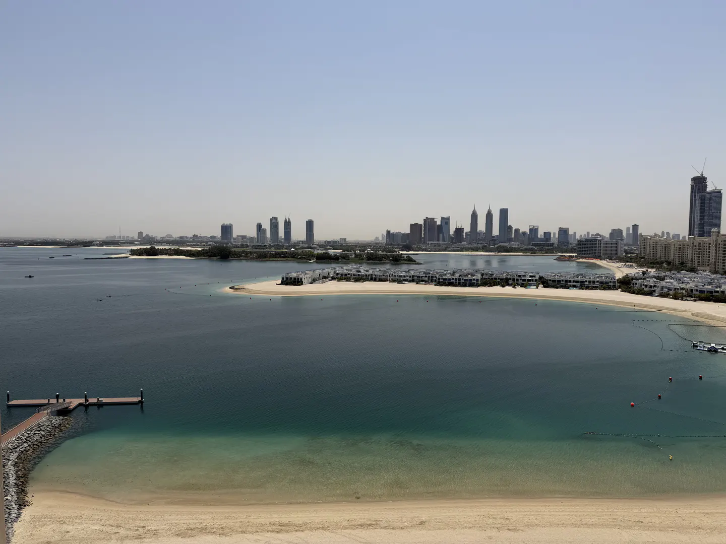 Dubai skyline view from a sandy beach with turquoise water. A pier extends into the water.