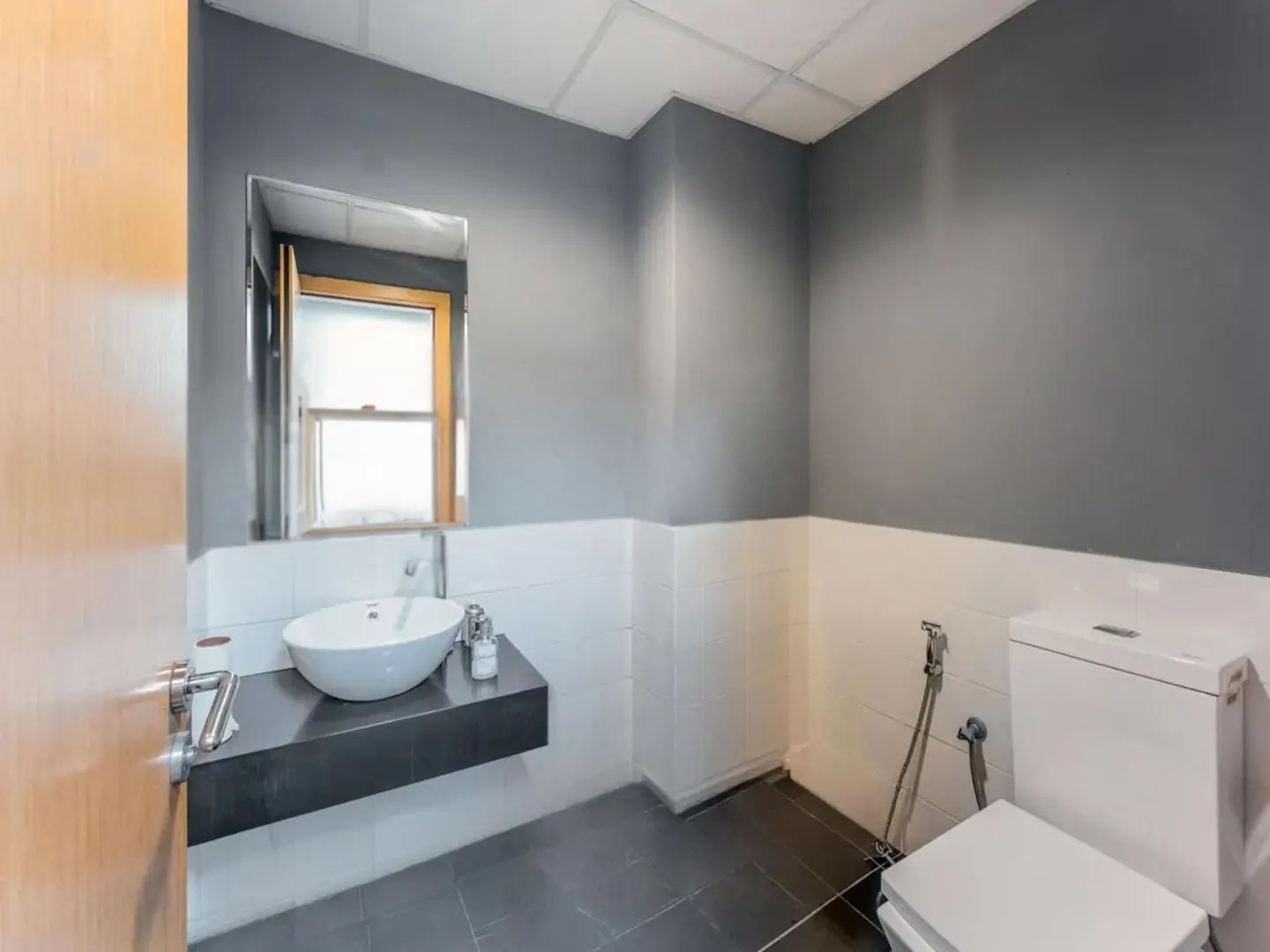 Bathroom with gray walls, white tile wainscoting, and dark gray tile floors. A white vessel sink sits on a black countertop. A toilet is visible in the corner.