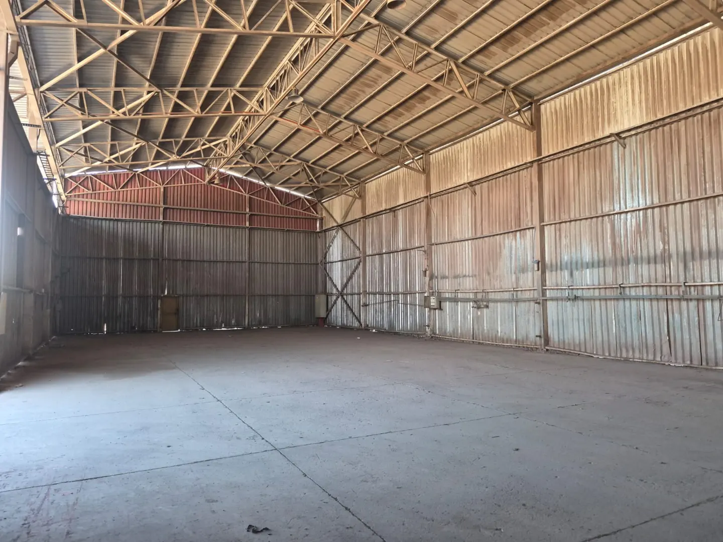 An expansive, empty industrial warehouse with high metal truss ceilings, corrugated metal walls, and a concrete floor.