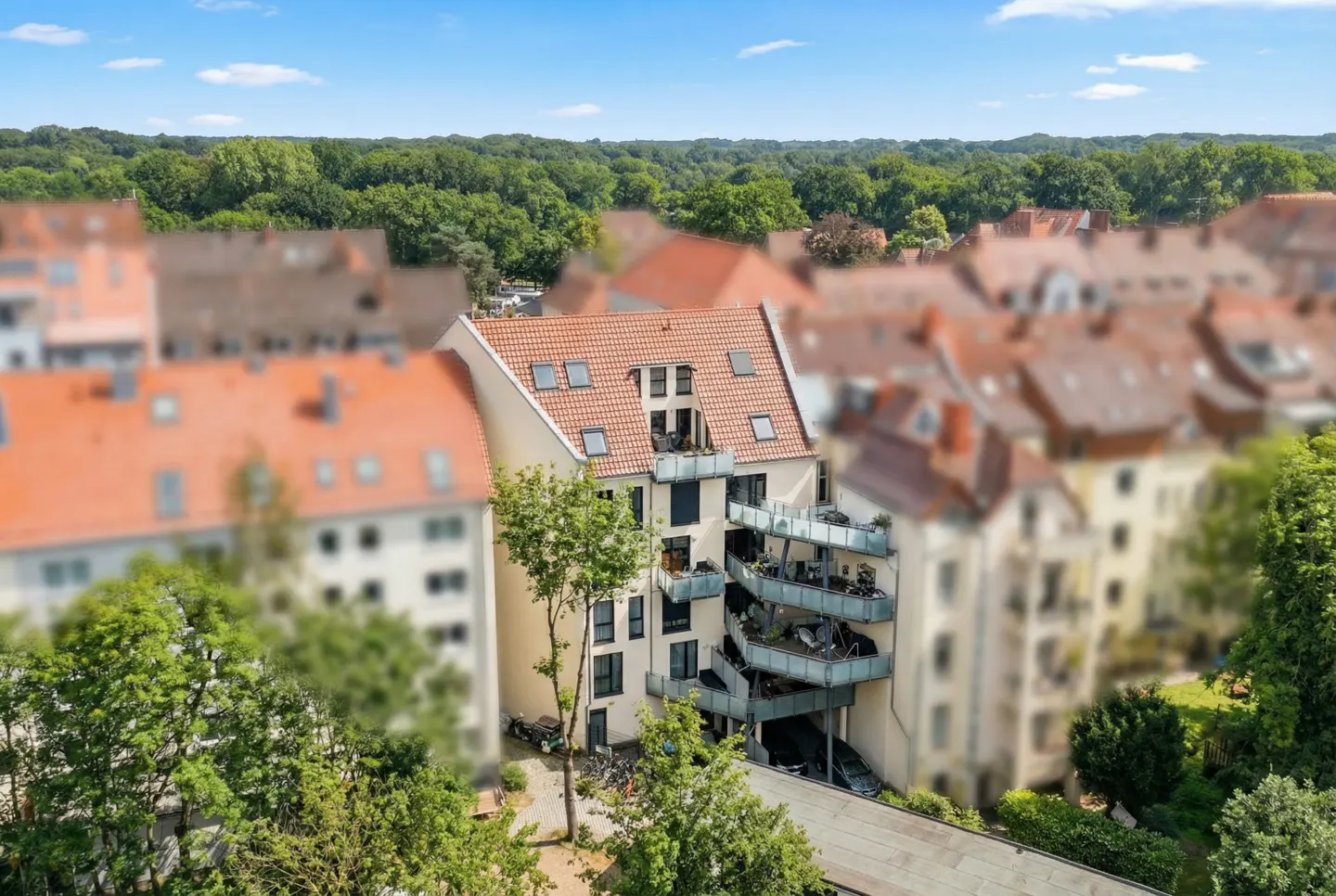 Aerial view of a modern apartment building with balconies, surrounded by trees and other buildings with red tile roofs.