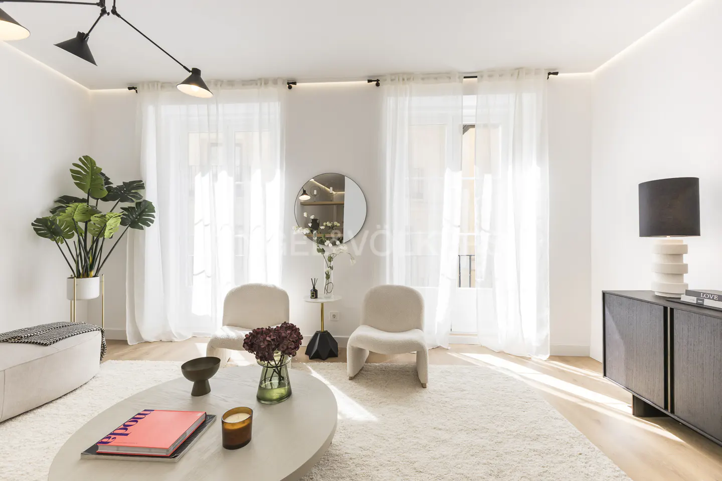 Bright living room with white walls, sheer curtains, and light wood floors. Two cream chairs face a round coffee table with books and flowers. A black cabinet and lamp are on the right.
