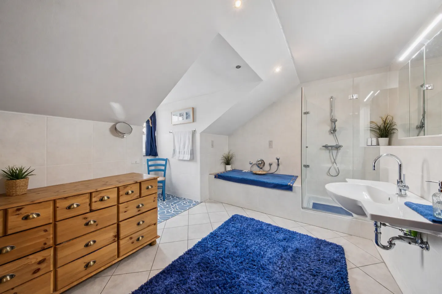 Bright bathroom with white walls, a wood dresser, and a blue rug. A glass shower and sink are on the right, and a blue chair sits by a window.