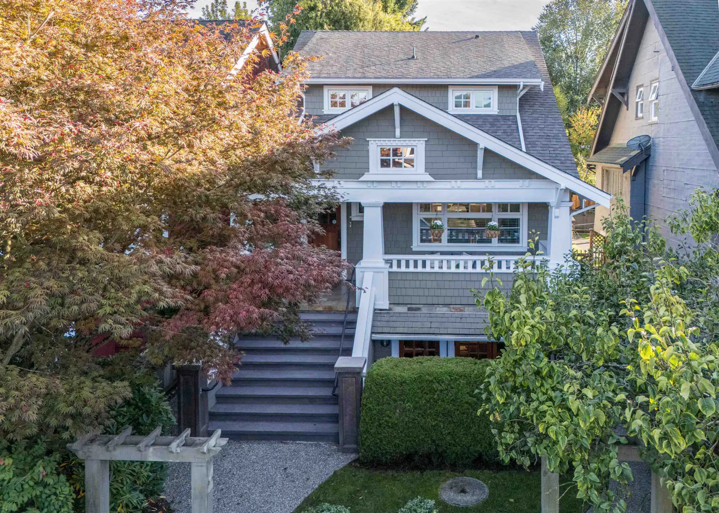 Exterior view of a two-story house with gray siding, white trim, and a front porch with steps. Trees and shrubs surround the house.