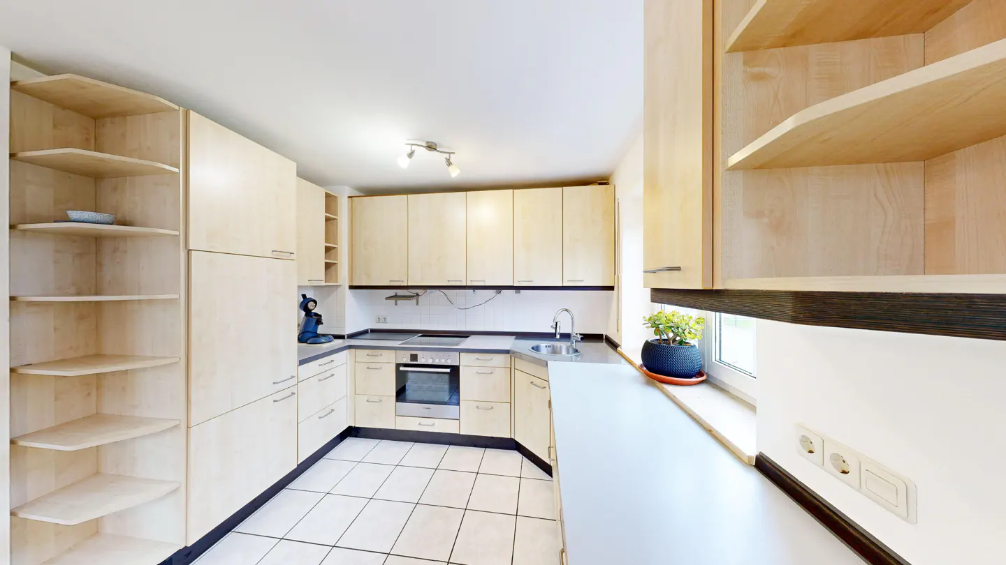 A bright kitchen with light wood cabinets, white tile floors, and a plant in a blue pot by the window.