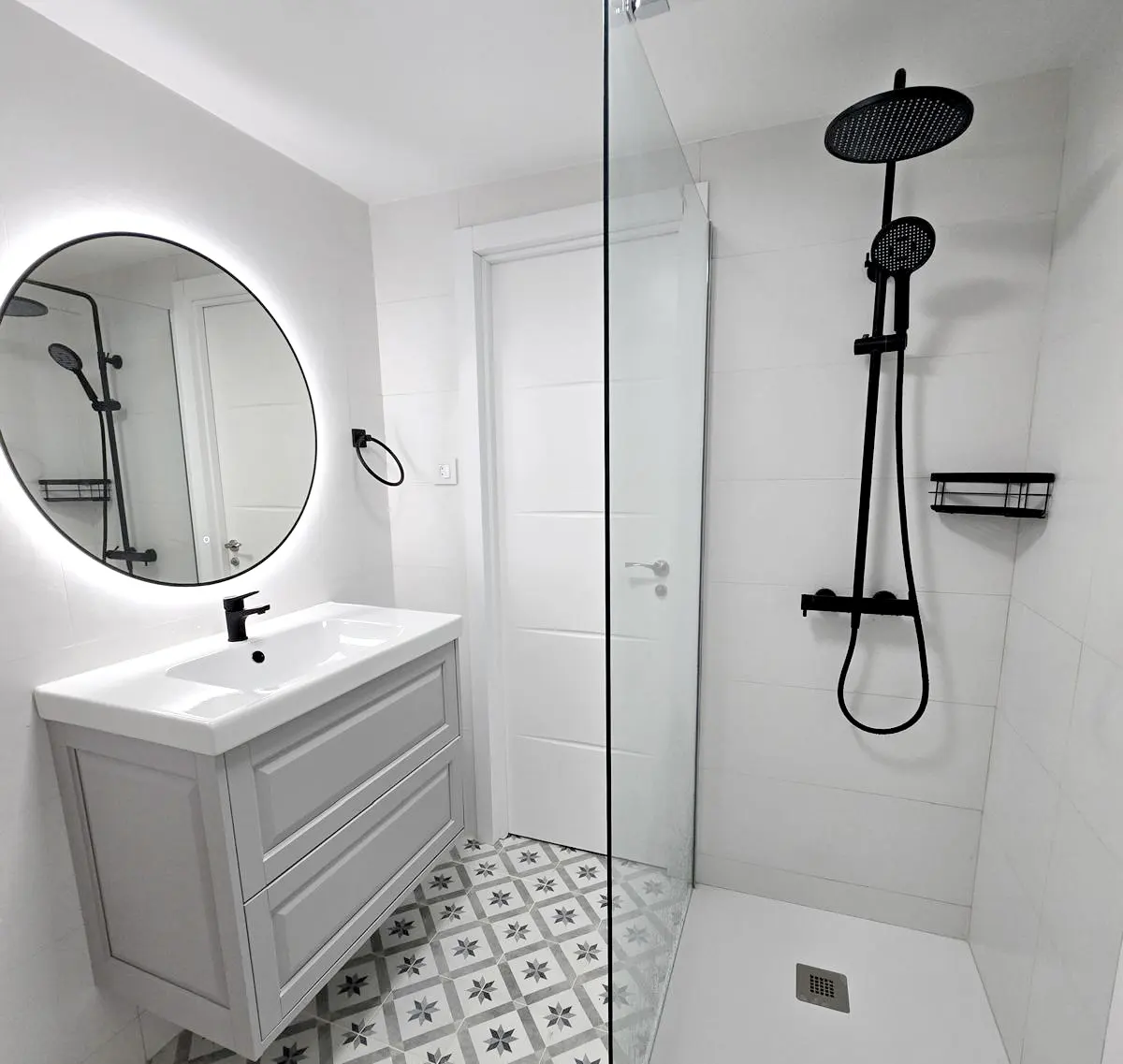 Modern bathroom with gray vanity, black fixtures, and a glass-enclosed shower with a black shower head. The floor has a patterned tile design.
