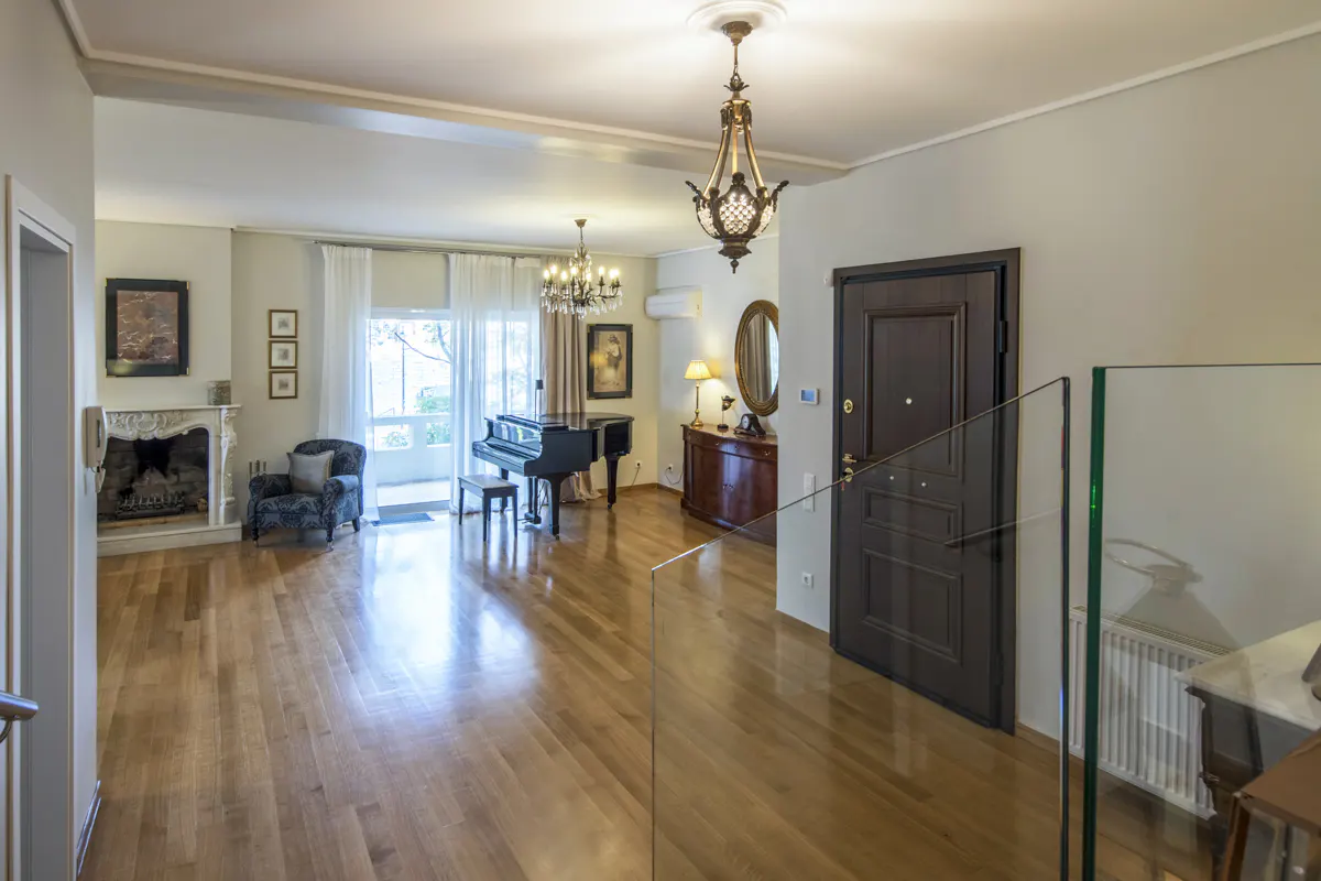 A bright living room with hardwood floors, a black piano, fireplace, and a dark wood front door.