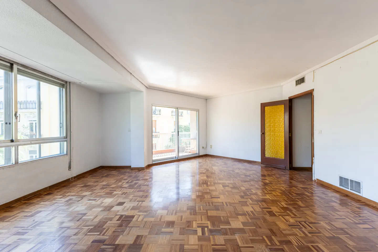 Bright, empty room with parquet floors, white walls, and large windows. A glass door leads to a balcony. Another door is open to a hallway.