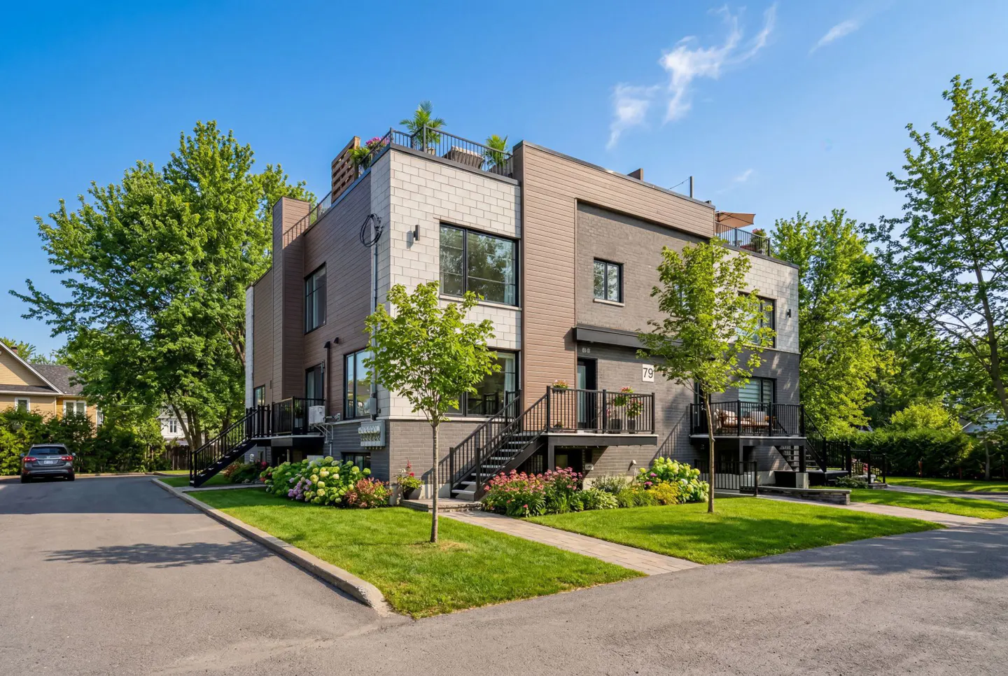 Modern townhouses with gray brick and wood siding, black railings, and rooftop patios, surrounded by green lawns and trees under a blue sky.