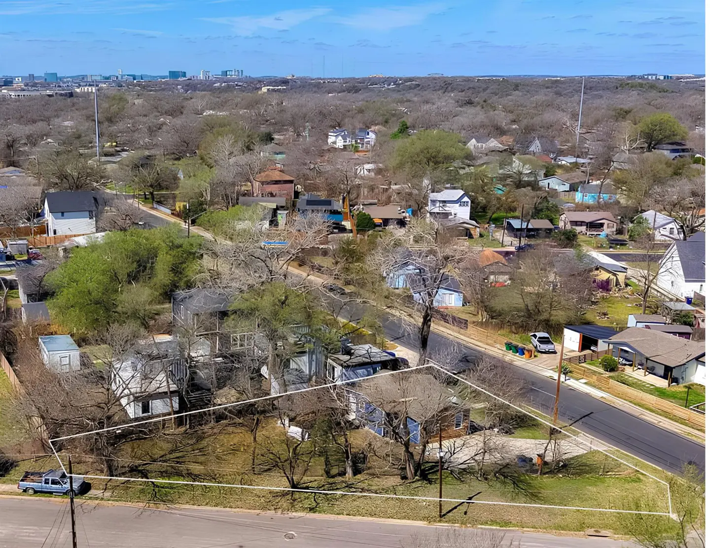 Aerial view of a residential lot outlined in white, with a blue house, trees, and a street. City skyline in the background under a blue sky.