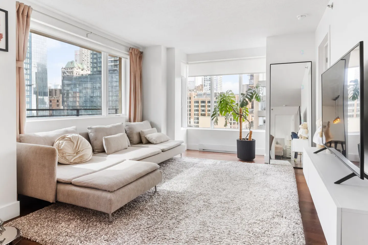 Bright living room with a beige sectional sofa, a large window overlooking city buildings, and a potted Monstera plant.