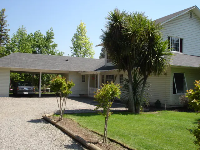 Two-story white house with a gray roof, a carport, and a gravel driveway. A green lawn and trees surround the house.