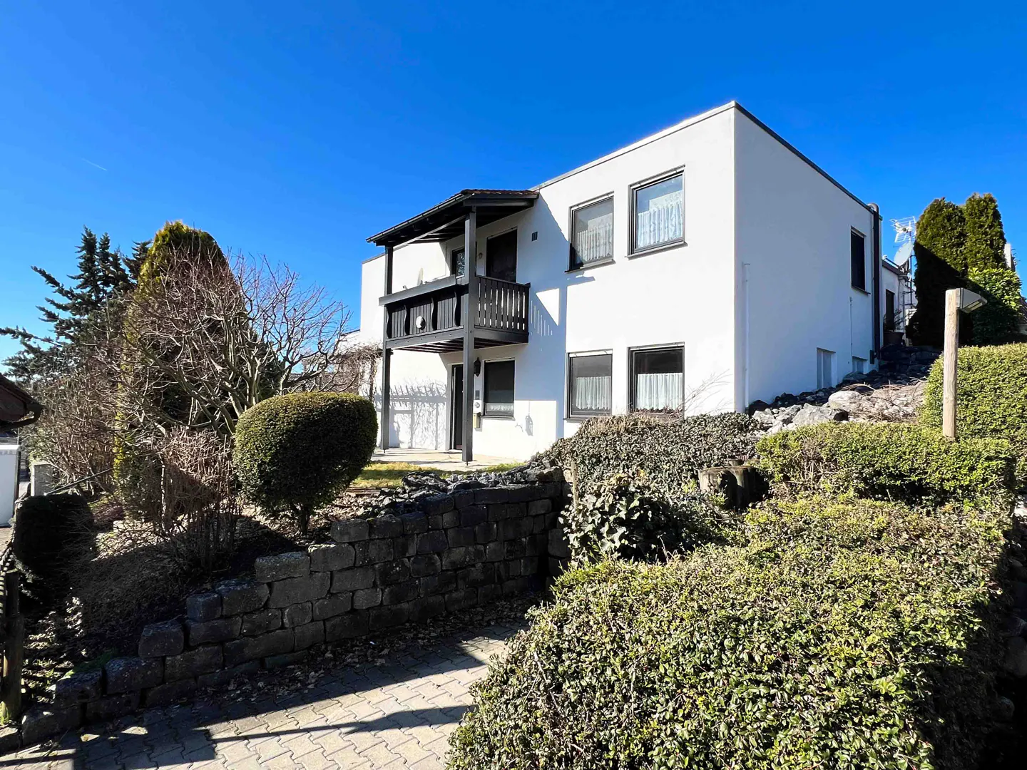 Two-story white house with a black balcony and dark-framed windows, set on a landscaped hill with stone walls and green bushes.
