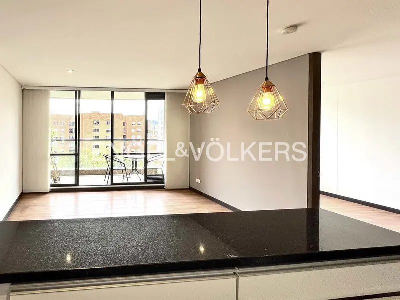 Modern apartment interior with wood floors, black granite countertop, and balcony with city view. Two geometric pendant lights hang above.