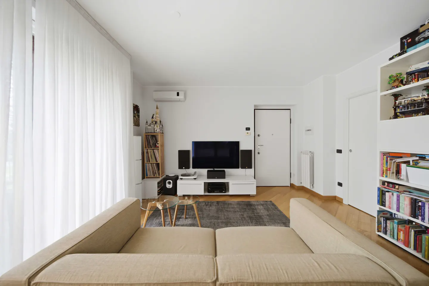 Bright living room with beige sofa, TV on white console, and white walls. Bookshelves and sheer curtains add to the airy feel.