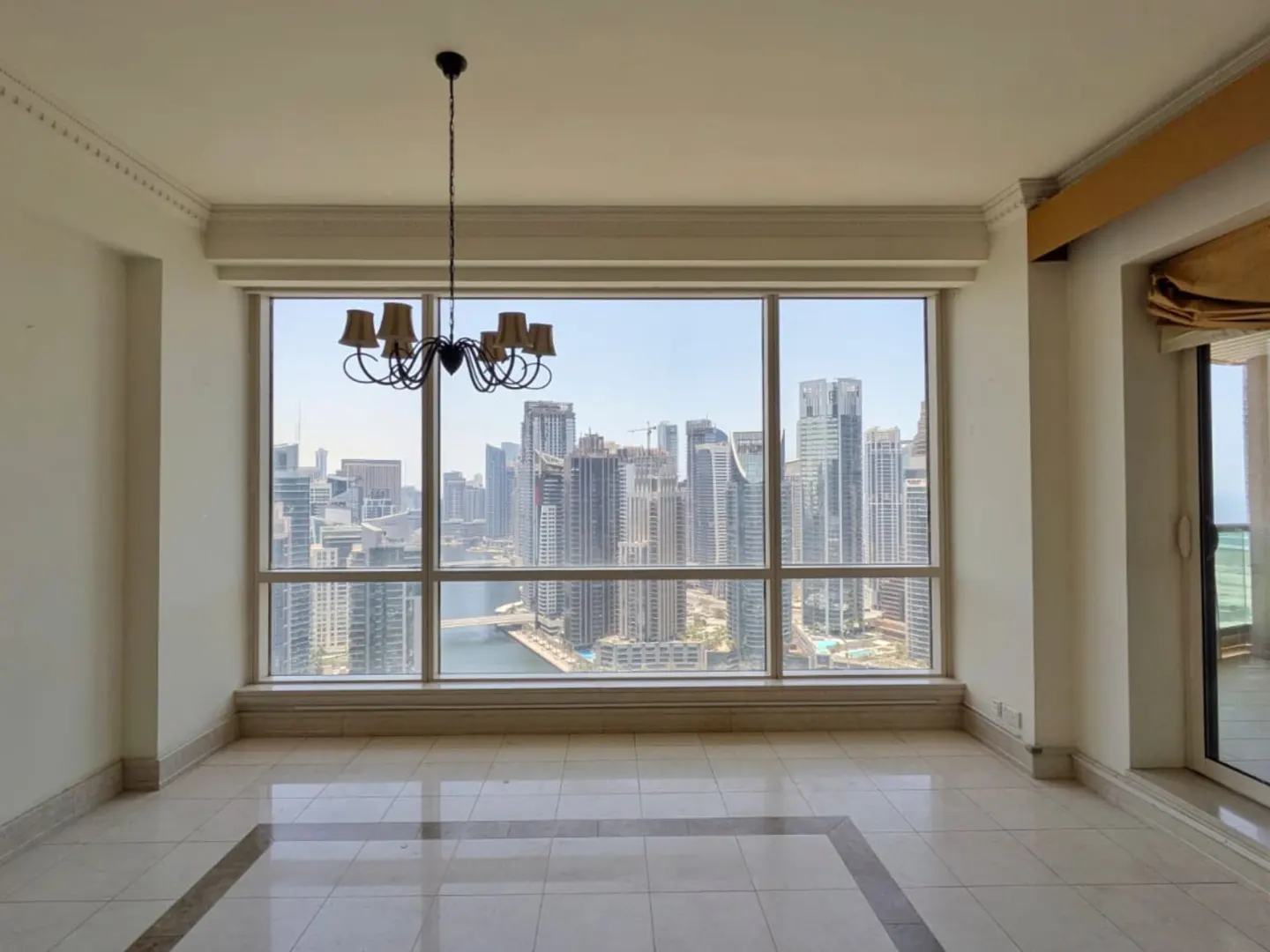 Empty room with white tile floors and a chandelier, featuring a large window overlooking a Dubai city skyline and canal.