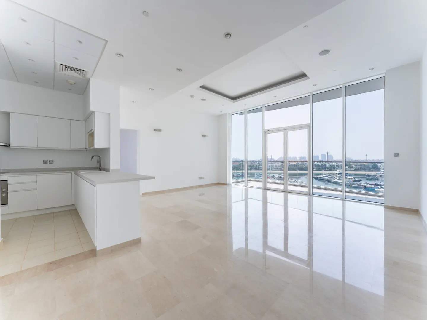 Bright, empty apartment with white walls, marble floors, and a kitchen. Large windows offer a city view.