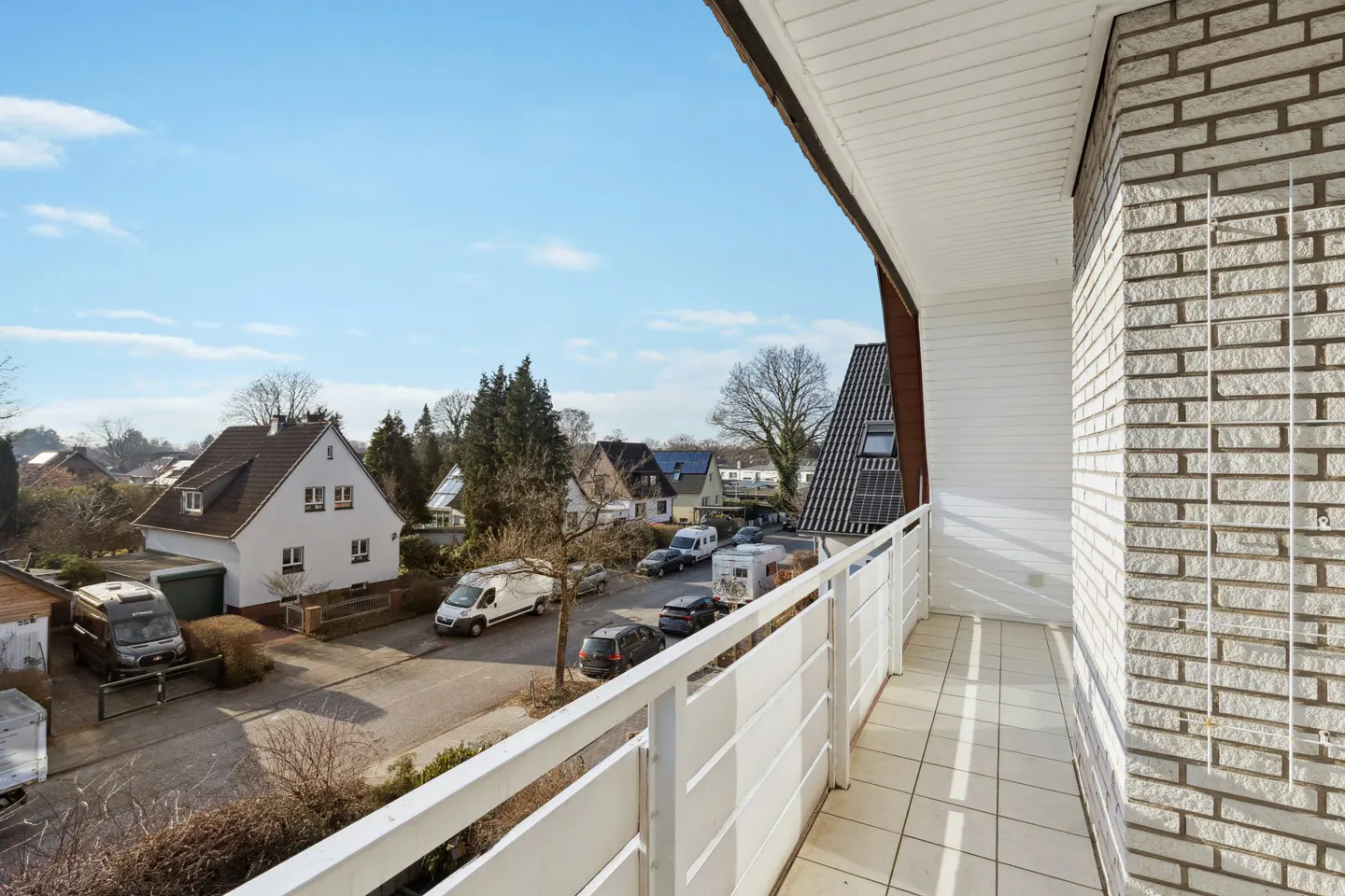 View from a balcony with white railings overlooking a street with houses and parked cars on a sunny day.