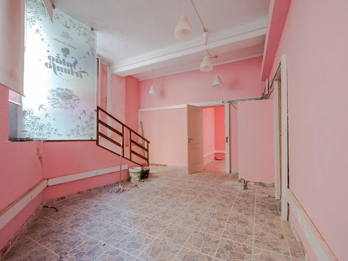 A pink room with tile floors, a staircase, and an open doorway. There are paint buckets and a mop in the corner.