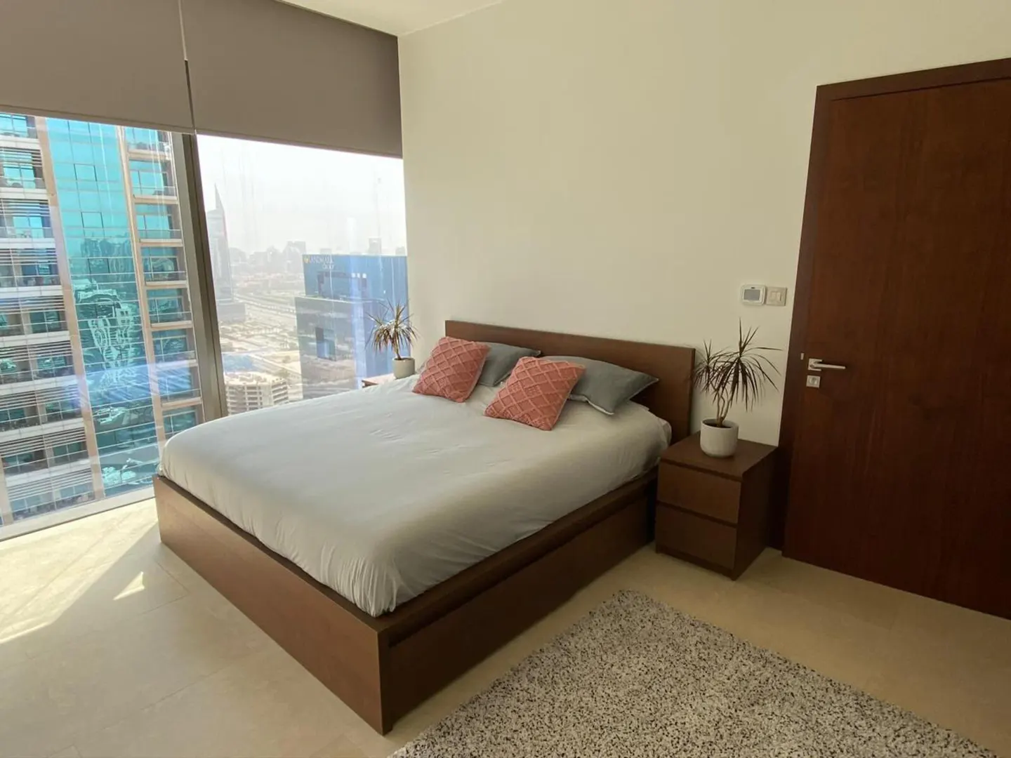 Modern bedroom with a queen bed, pink accent pillows, wooden furniture, and floor-to-ceiling windows with city views.