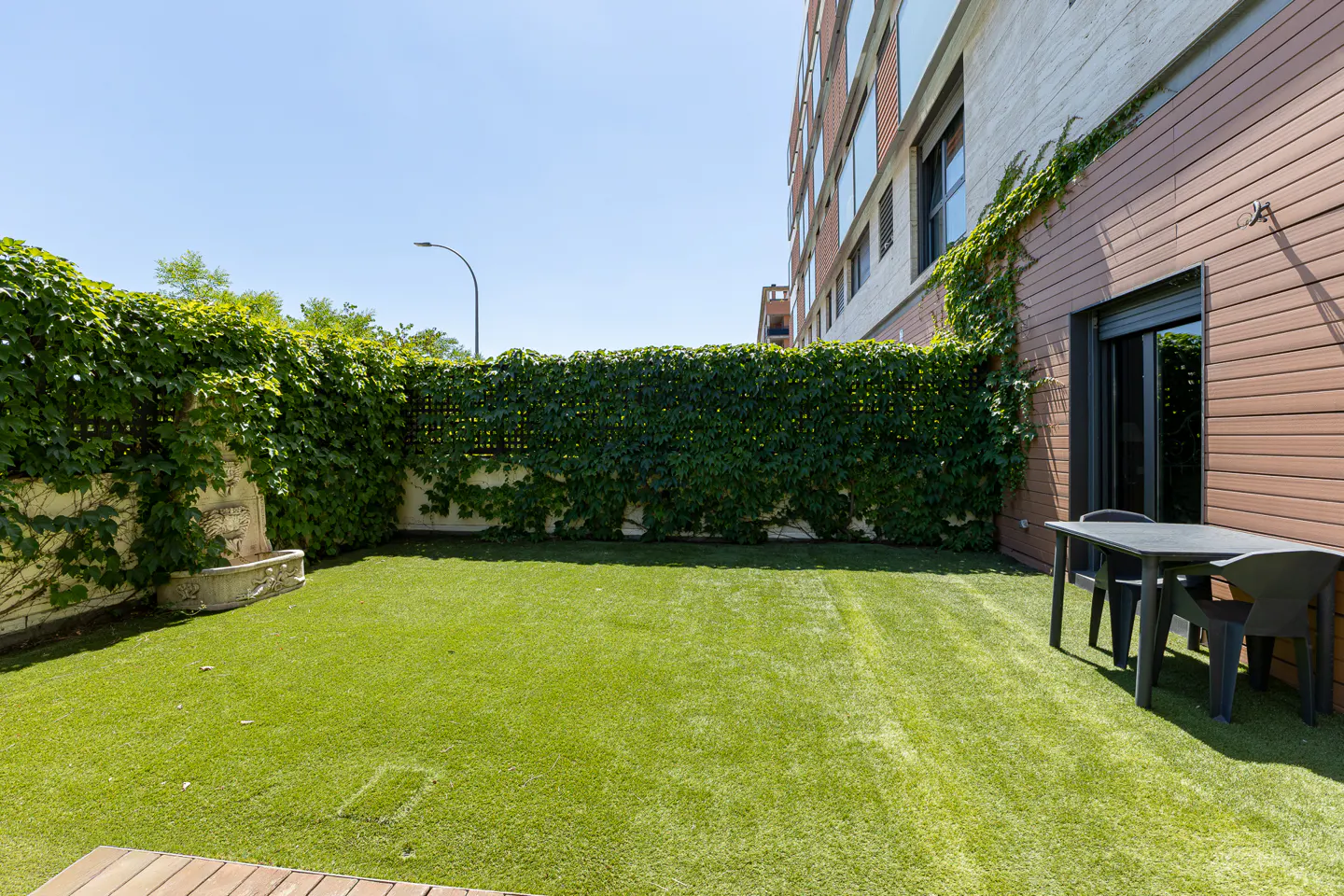 A private backyard with a green lawn, a tall ivy-covered fence, a stone fountain, and a small table with chairs.