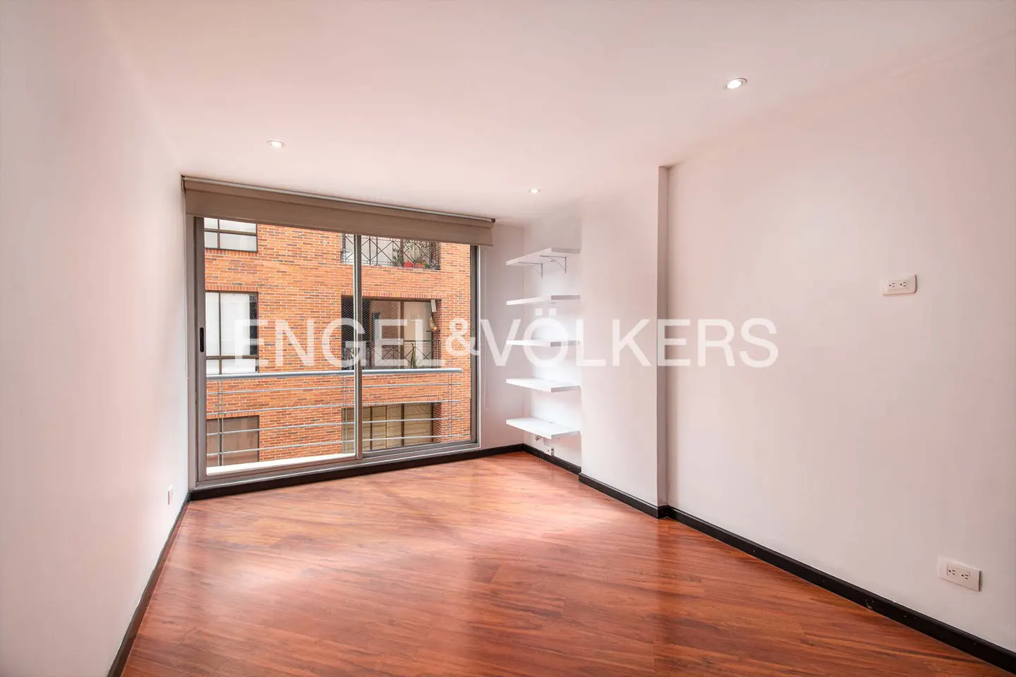 Empty room with hardwood floors, white walls, and a large window showing a brick building. White shelves are on the wall.