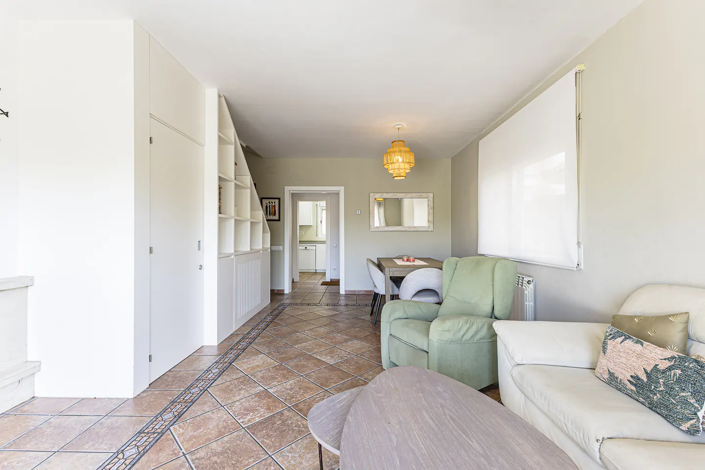 Living room with white sofa, green armchair, and dining table. White built-in shelves and brown tiled floor.
