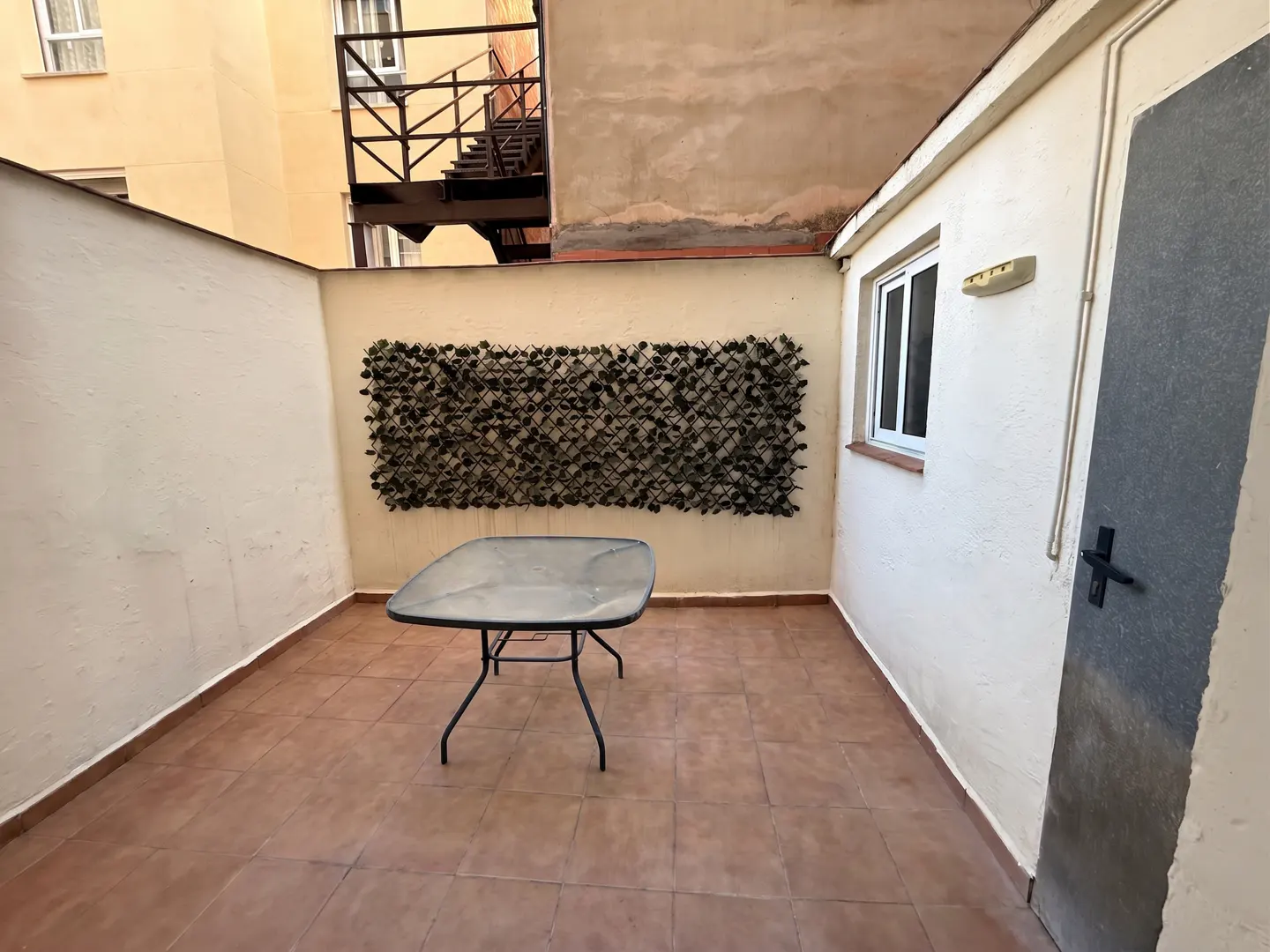 A small outdoor patio with a glass table, tiled floor, and a decorative green wall panel. A window and door are visible on the right.
