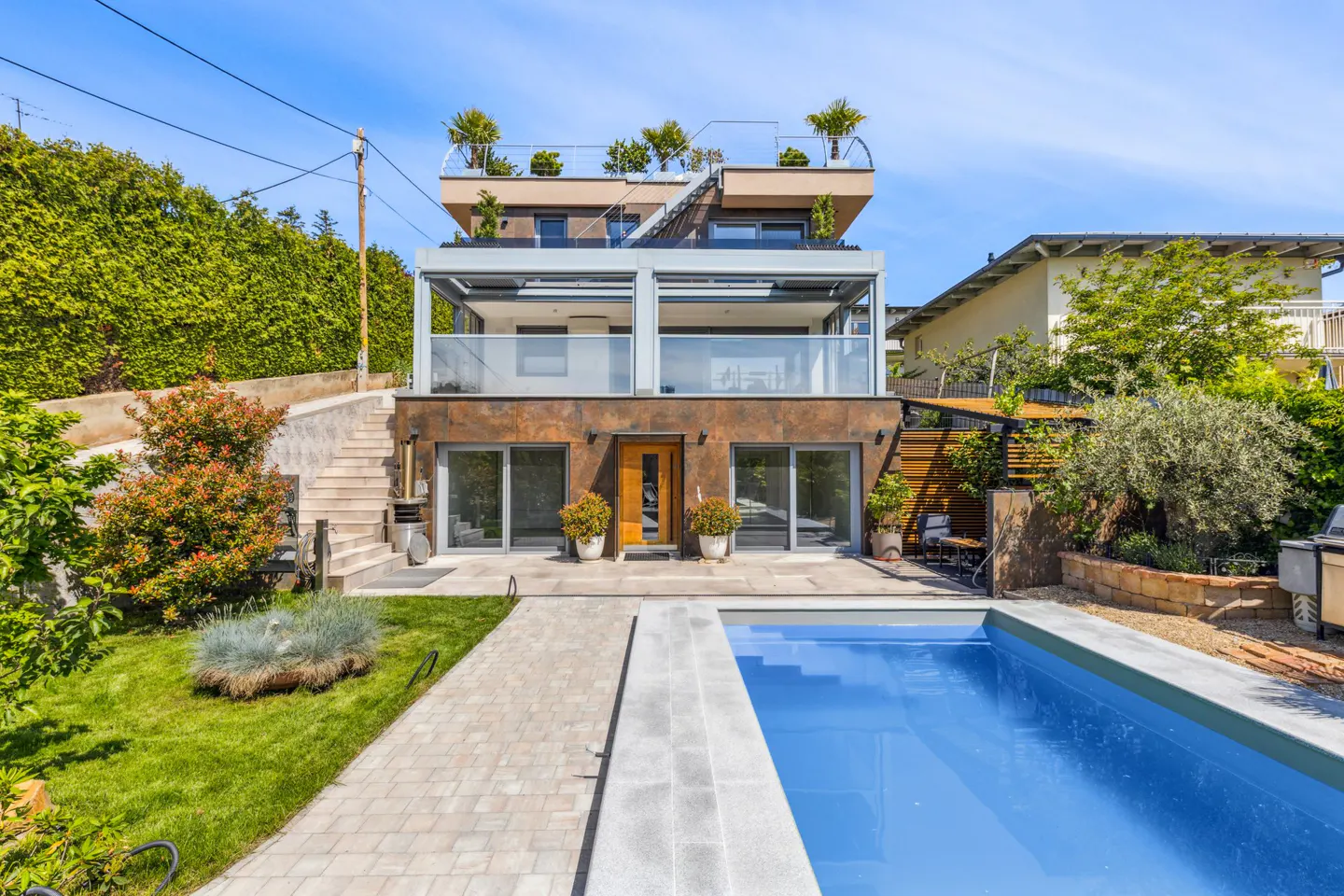 Modern three-story home with a rooftop terrace, glass balconies, stone facade, and a backyard swimming pool on a sunny day.