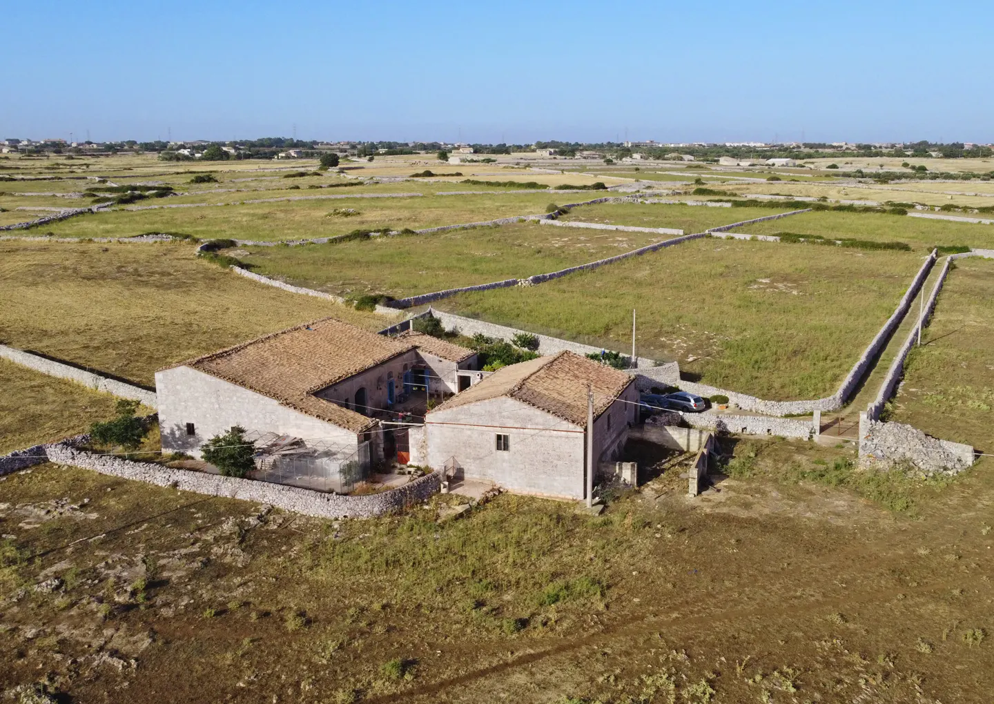 Aerial view of a stone farmhouse with a tile roof, surrounded by fields enclosed by stone walls under a clear blue sky.