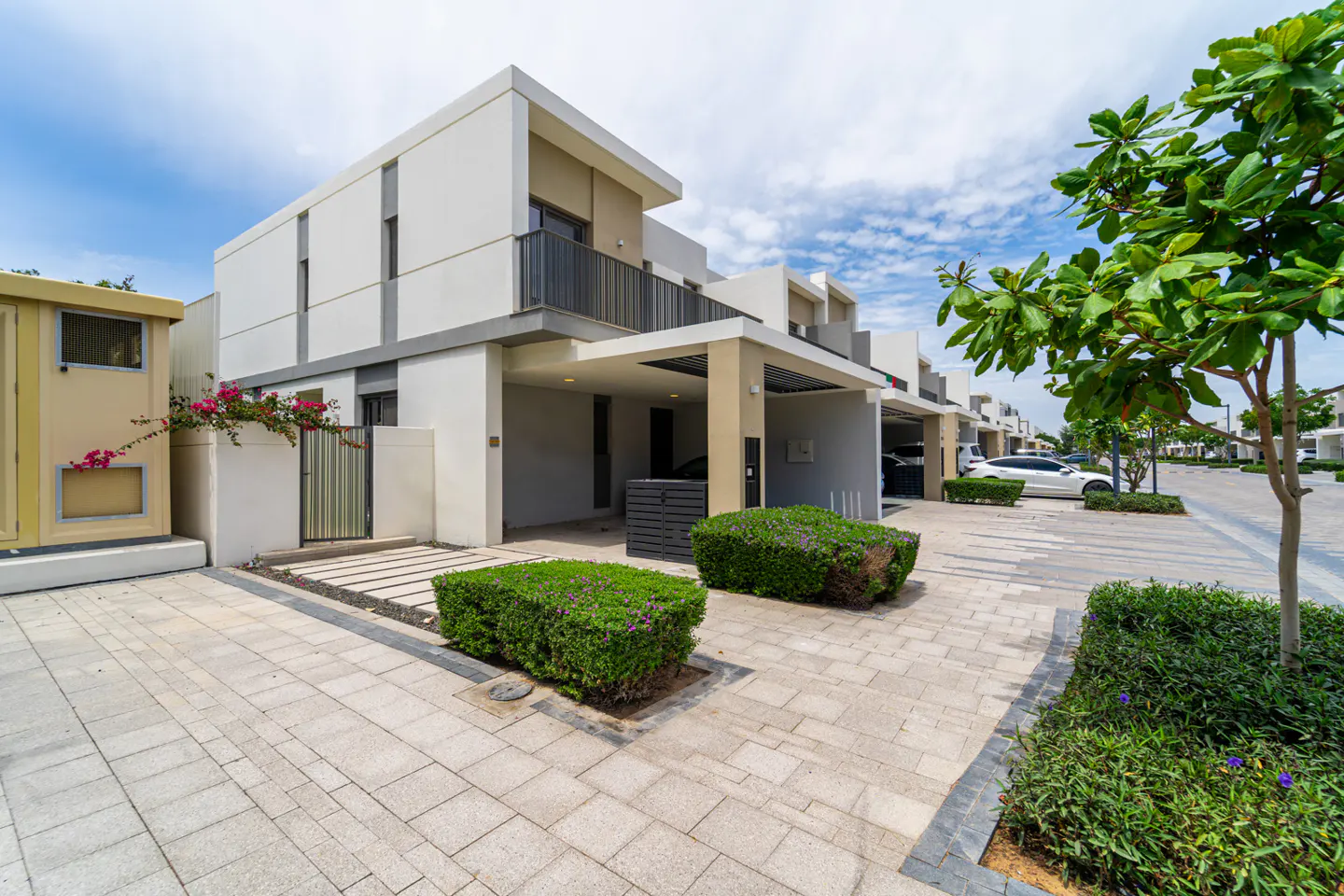 Modern two-story townhouse with a carport and balcony. The exterior is white with gray accents, and there are green bushes in front.