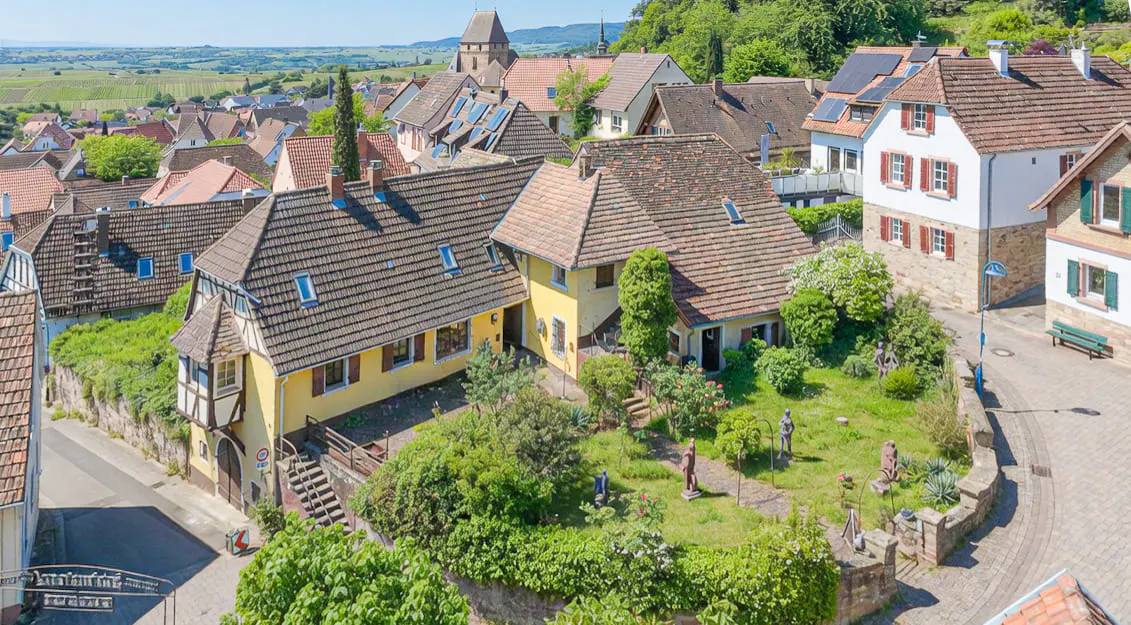 Aerial view of a European village with yellow houses, red tile roofs, and green gardens on a sunny day.