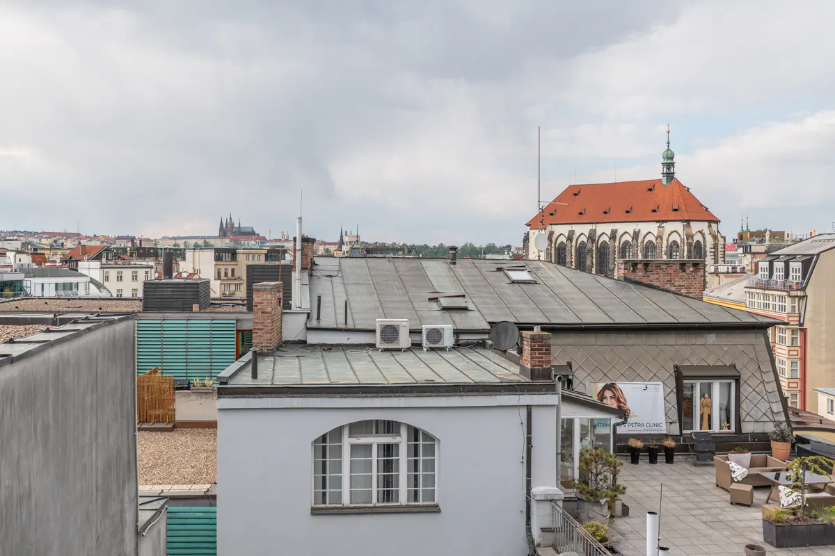 Rooftop view of Prague cityscape with buildings, church with red roof, and outdoor terrace with furniture. Cloudy sky.