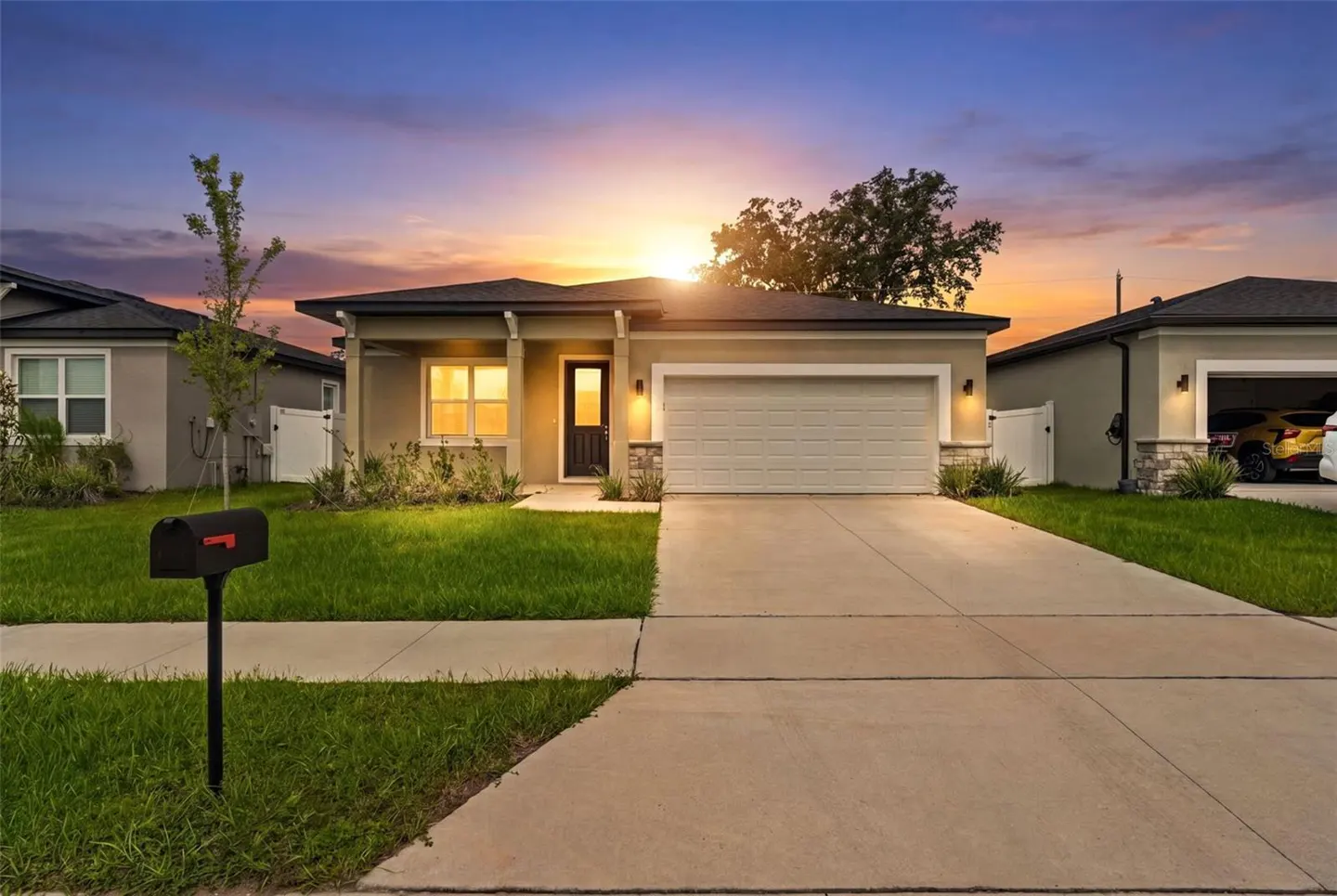 Single-story suburban home with a concrete driveway, green lawn, and mailbox at sunset with a colorful sky.