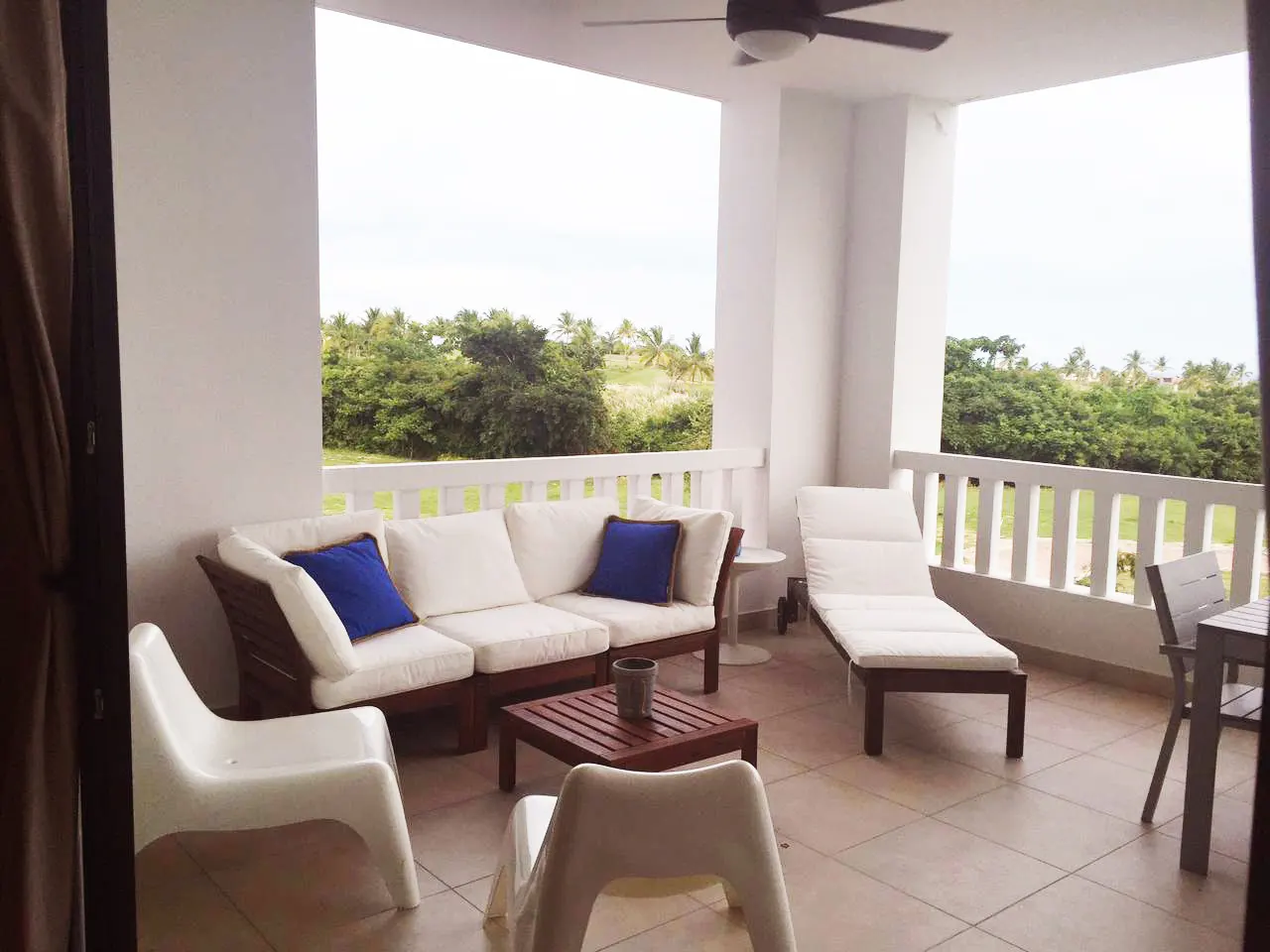 Balcony with white sofa, blue pillows, wood table, lounge chair, and white chairs. Green trees and white railing in background.