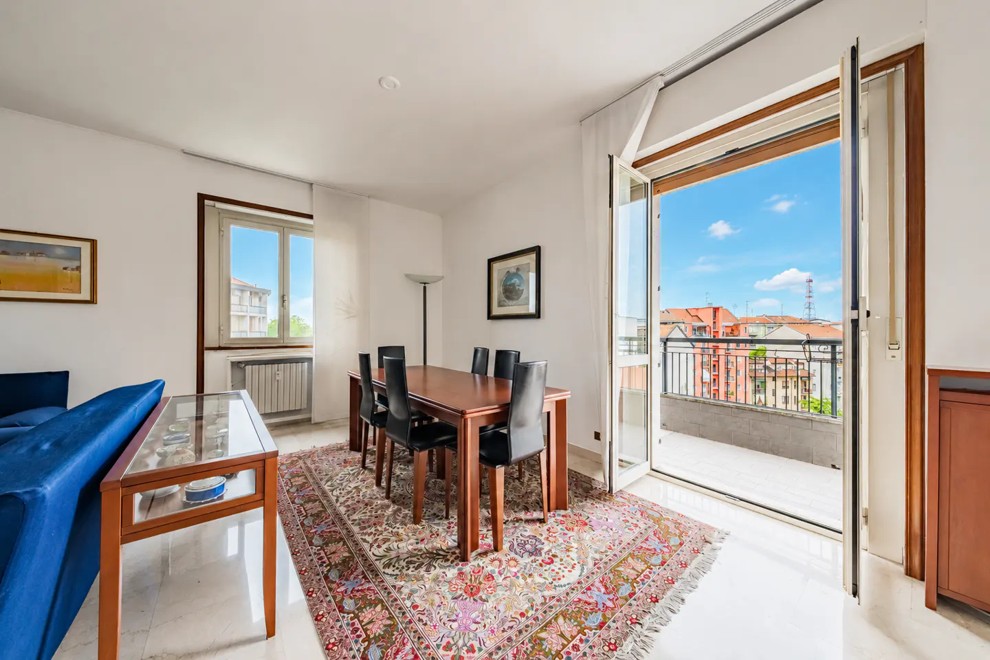 Bright dining room with a wood table, black chairs, and a colorful rug. Balcony doors open to a city view under a blue sky.