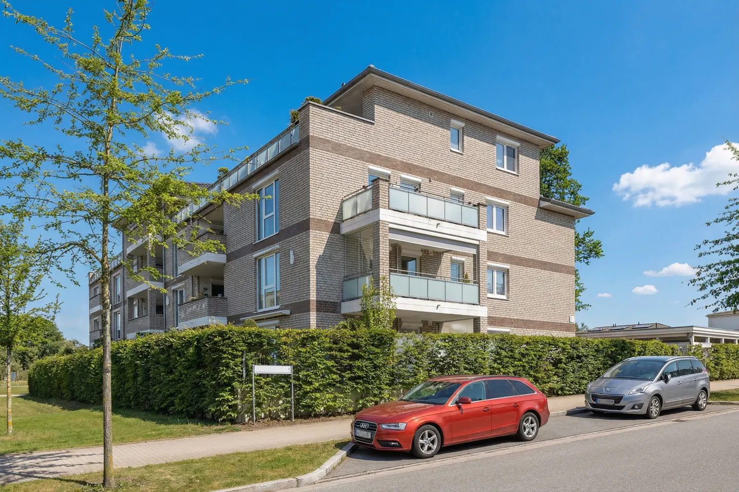 Three-story brick apartment building with balconies, green hedge, and two parked cars, one red and one silver, under a blue sky.