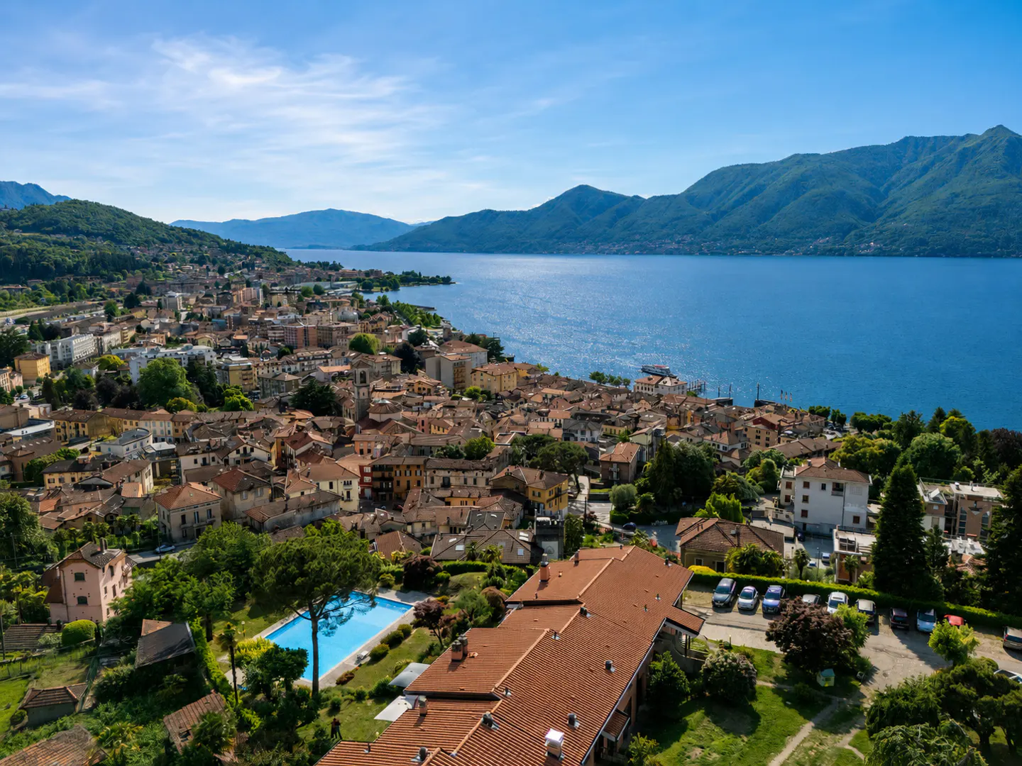 Aerial view of a coastal town with red-roofed buildings, a blue pool, and a lake with mountains in the background under a clear blue sky.