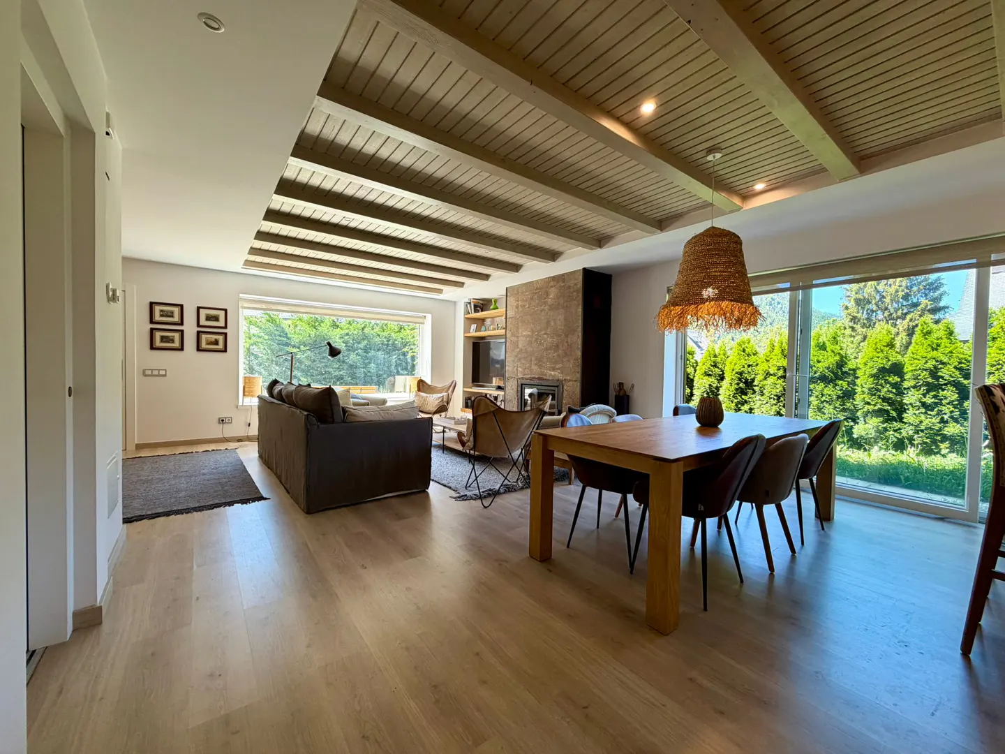 Open-plan living and dining area with light wood floors, beamed ceiling, stone fireplace, and large windows to a garden.