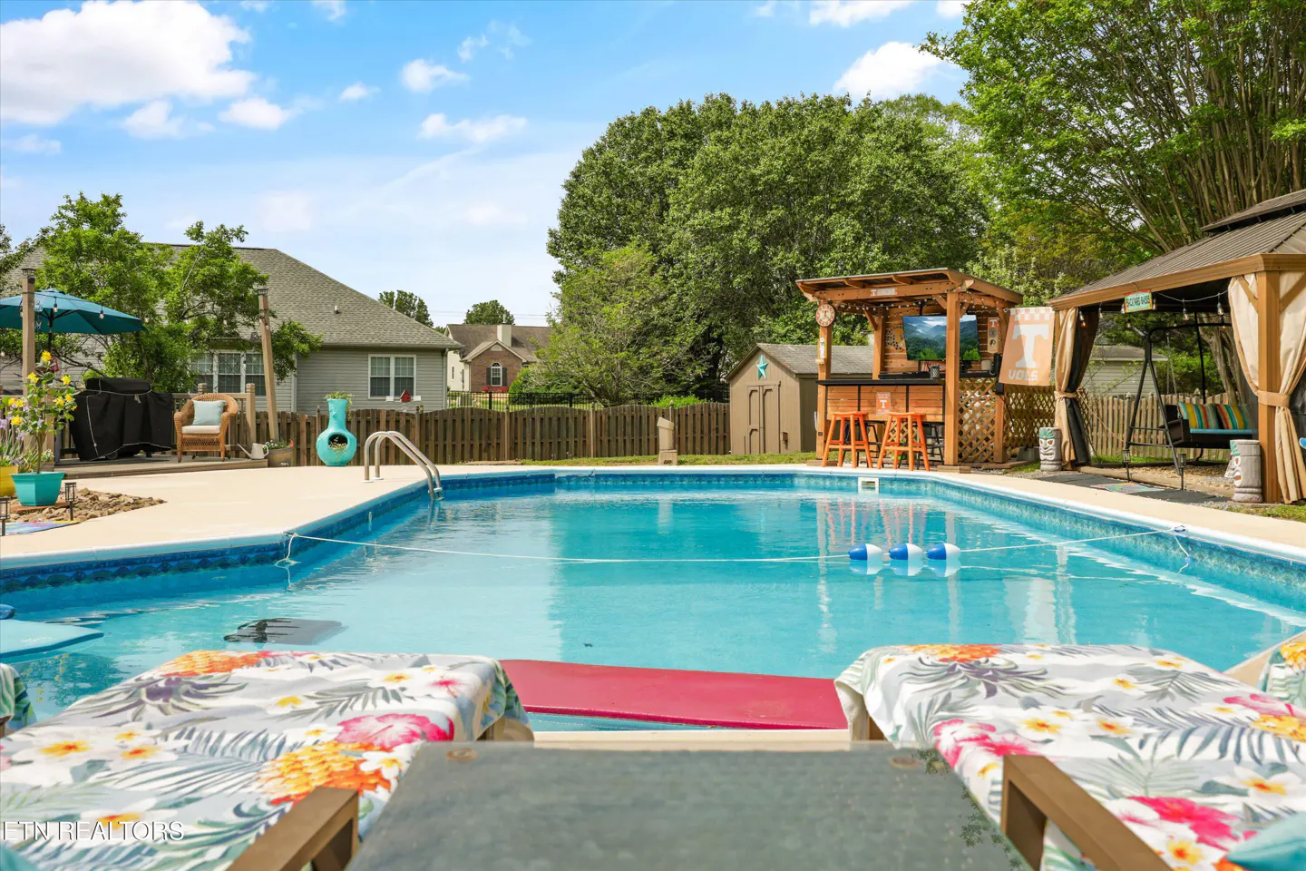 Backyard pool with lounge chairs, bar, and gazebo. Blue water reflects the sky. Trees and houses are in the background.