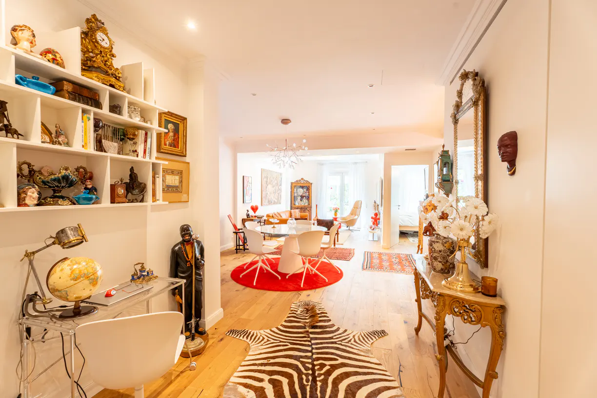 Bright, eclectic home interior with a zebra rug, red circular rug, and a desk with a globe. A white bookshelf displays various objects.