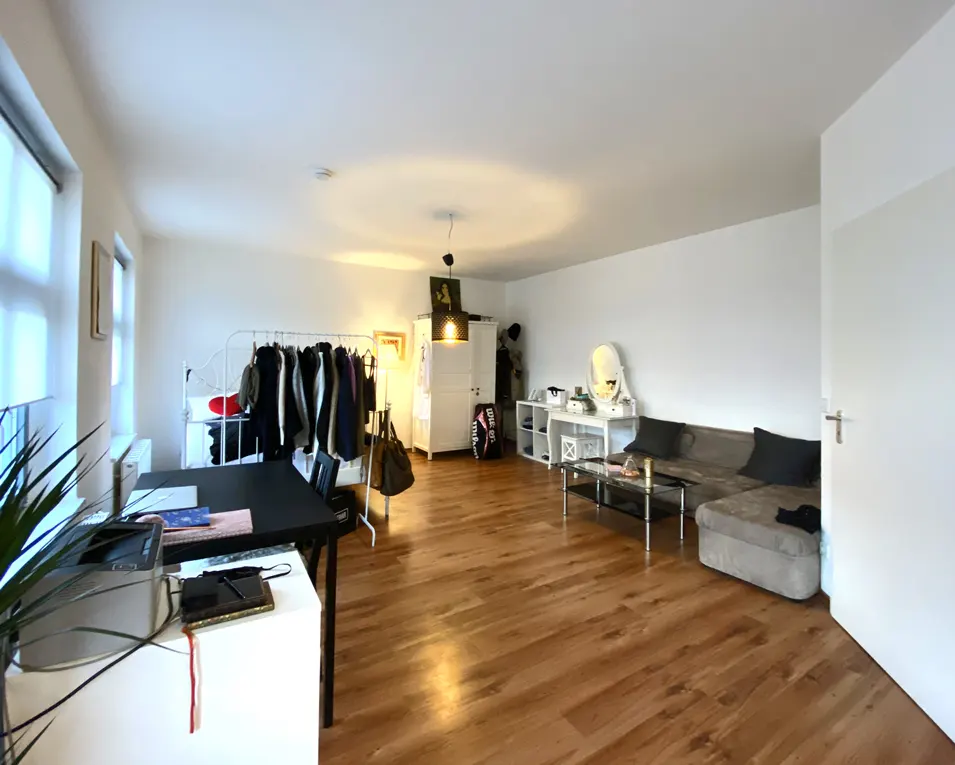 Bright, open apartment with wood floors. A clothing rack, desk, sofa, and vanity are visible. The walls are white.